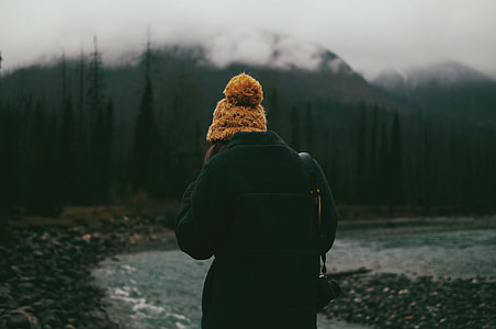 person wearing black bubble jacket walking near grey body of water and trees during daytime thumbnail