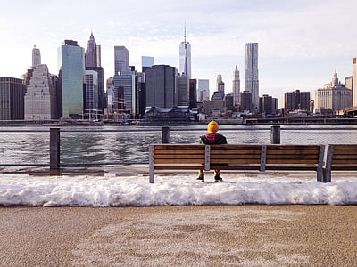 person sitting on brown wooden bench on baywalk overlooking inline high rise buildings across the body of water at daytime thumbnail