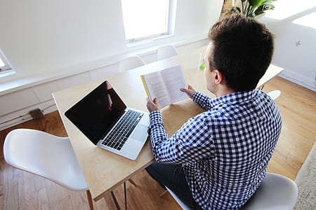 man reading book near laptop while leaning on table thumbnail