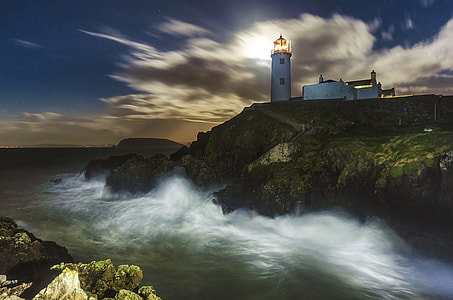 time lapse photography of waves hitting cliff with lighthouse during daytime thumbnail