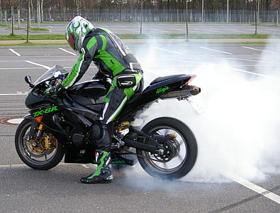 person wearing black and green racing suit performing rear tire burnout on black Kawasaki Ninja ZX-6r on empty parking lot during daytime thumbnail