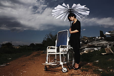 woman standing beside empty wheelchair holding broken umbrella thumbnail