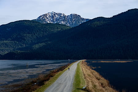 person walking on footpath between body of water in front of mountain at daytime thumbnail