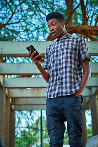 man wearing white, black, and gray gingham button-up collared t-shirt holding black smartphone during daytime thumbnail