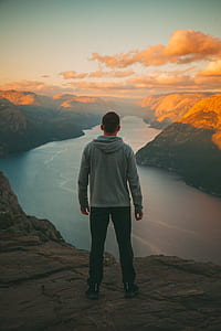 man standing on cliff facing body of water during golden hour thumbnail