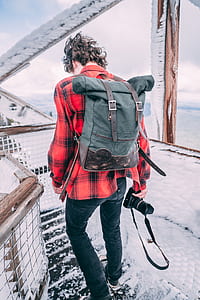 man with green knapsack carrying black DSLR camera while walking on snow-covered wooden bridge thumbnail