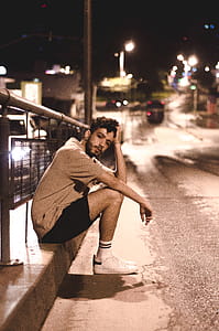 Man Wears Brown Shirt and Black Shorts Seats Beside Black Street Post thumbnail