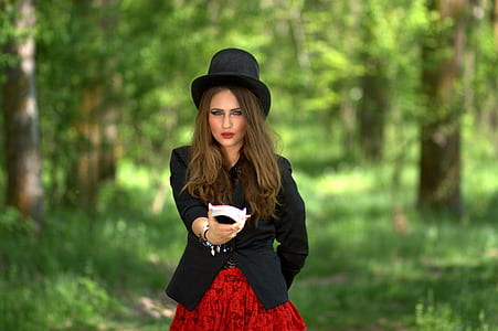 shallow focus photography of woman wearing black formal coat and hat standing in the middle of trees during daytime thumbnail