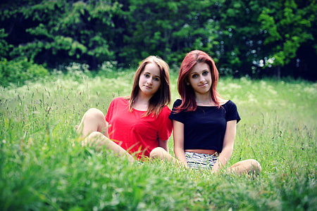 two woman in red and black seated in the middle of green grass field at daytime thumbnail