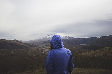 person wearing blue hooded jacket standing on elevated surface facing mountains during daytime thumbnail