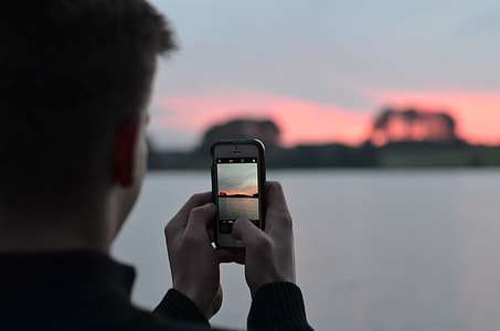 man holding smartphone white taking a photo of sunset beside body of water thumbnail