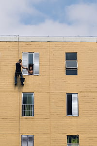 man hanging on rope with bucket beside person at the window inside building during day thumbnail
