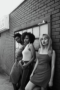 grayscale photo of three standing woman with brick wall behind them thumbnail