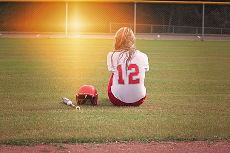 woman in white and red 12 jersey sitting on the field beside baseball bat and batting hat thumbnail