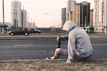 man wearing hoodie sitting beside the road holding bottle near cars during day thumbnail