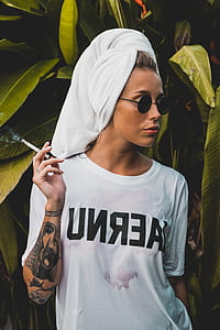 woman wearing towel and white crew-neck t-shirt holding cigarette standing near green leaf plant during daytime thumbnail