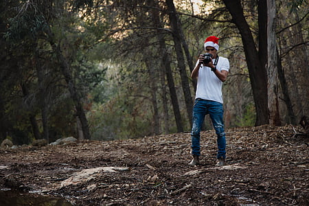 man in white t-shirt and blue distressed jeans holding DSLR camera in forest during daytime thumbnail