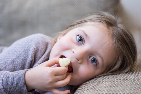 girl wearing gray long-sleeved shirt lying on brown fabric sofa while eating thumbnail
