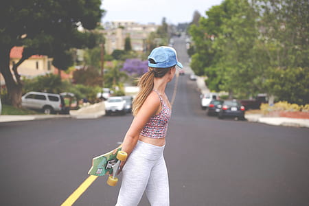 selective focus photography of woman wearing pink multicolored spaghetti strap on road holding skateboard thumbnail