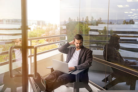 man sitting on gray wooden Adirondack chair using gray laptop near glass wall thumbnail