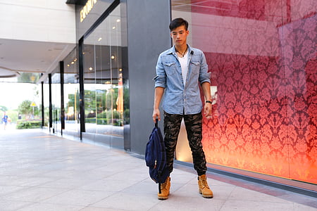 standing man wearing blue chambray long-sleeved shirt, camouflage pants and tan boots holding blue bag thumbnail