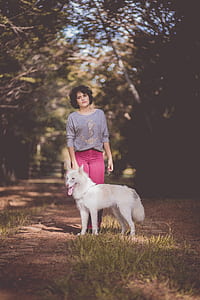 Woman in Purple Long Sleeve Shirt and Pink Jeans Standing Next to White German Spitz With Background of Green Trees during Day thumbnail