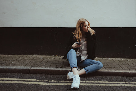 woman wearing black cardigan and blue denim jeans sitting beside black concrete road holding black smartphone thumbnail