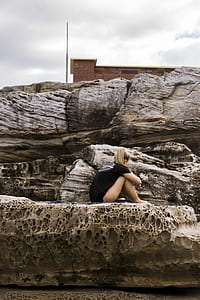 woman wearing black t-shirt sitting on black stone formation on beach during daytime thumbnail