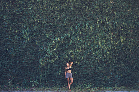 photo of woman wearing black bikini top and gray shorts standing near plant wall thumbnail