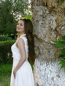 woman in white sleeveless dress looking up beside brown tree trunk thumbnail