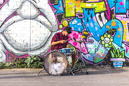 man wearing black and red checkered long-sleeved collared shirt playing drums near graffiti painted wall during daytime thumbnail