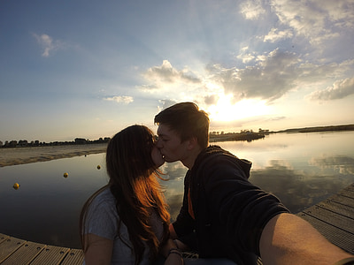 panoramic photography of man wearing black jacket kissing woman wearing gray t-shirt standing near body of water thumbnail