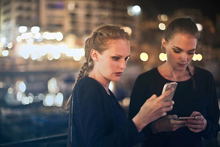 Two young blonde women in black dresses texting on their mobile phones at night outdoors thumbnail