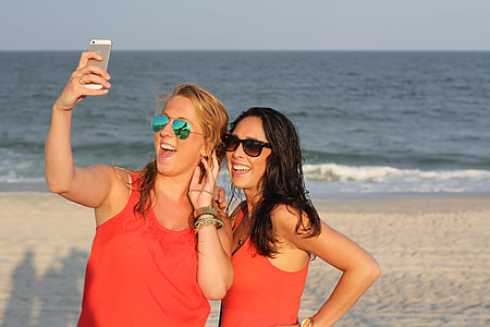 two woman wearing red tank tops taking photos on white sand beach shore thumbnail