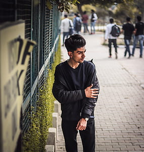 man wearing black leather jacket standing on sidewalk beside green metal wall near people beside trees during daytime thumbnail