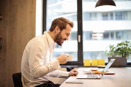 A young bearded caucasian man in formals looking at his laptop screen while holding a cup thumbnail