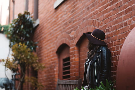 person wearing black jacket and black cap leaning on brown brick wall thumbnail