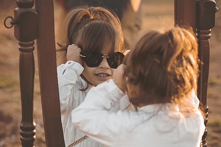 Girl in White Long-sleeved Shirt Wearing Sunglasses Facing Mirror thumbnail
