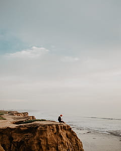 man sitting on the cliff edge under gray sky at daytime thumbnail