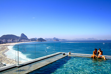 couple near corner of swimming pool on high rise building with ocean view with people on shore under blue-sky during daytime \ thumbnail