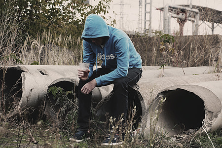 man wearing black pants and blue Adidas pullover hoodie sitting on gray concrete poles holding clear disposable cup thumbnail