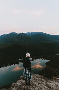woman standing on cliff facing body of water and mountain thumbnail