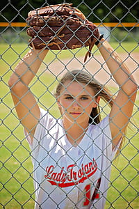 woman wearing white and red Lady Trojant button-up jersey and brown baseball mitt leaning on gray chain link fence thumbnail