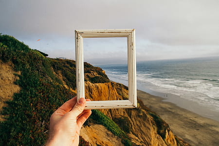 person holding white wooden photo frame facing on seashore and mountain during daytime thumbnail
