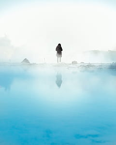 person standing on snow covered ground beside body of water surrounded by fogs thumbnail