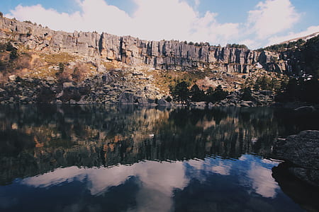 Grey Rocky Mountain Near Body of Water Under Blue Sky With White Clouds during Day Time thumbnail