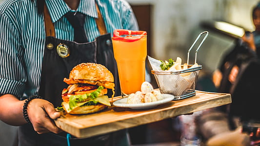 person wearing black apron carrying brown wooden serving tray with burger and glass of orange juicer thumbnail