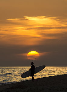 silhouette photo of man standing on seashore while holding surfboard during golden hour thumbnail