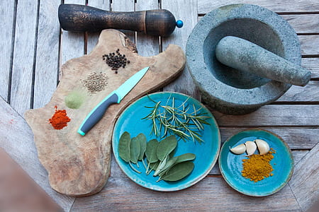 gray mortar and pestle next to blue saucer with garlic and brown powder next to blue plate with green leafs next to brown chopping board with knife and four seasoning powders thumbnail