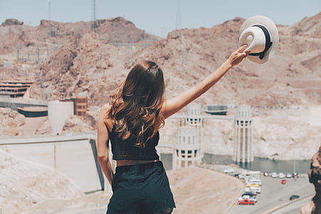 woman wearing black outfit holding white fedora hat facing rock formation thumbnail